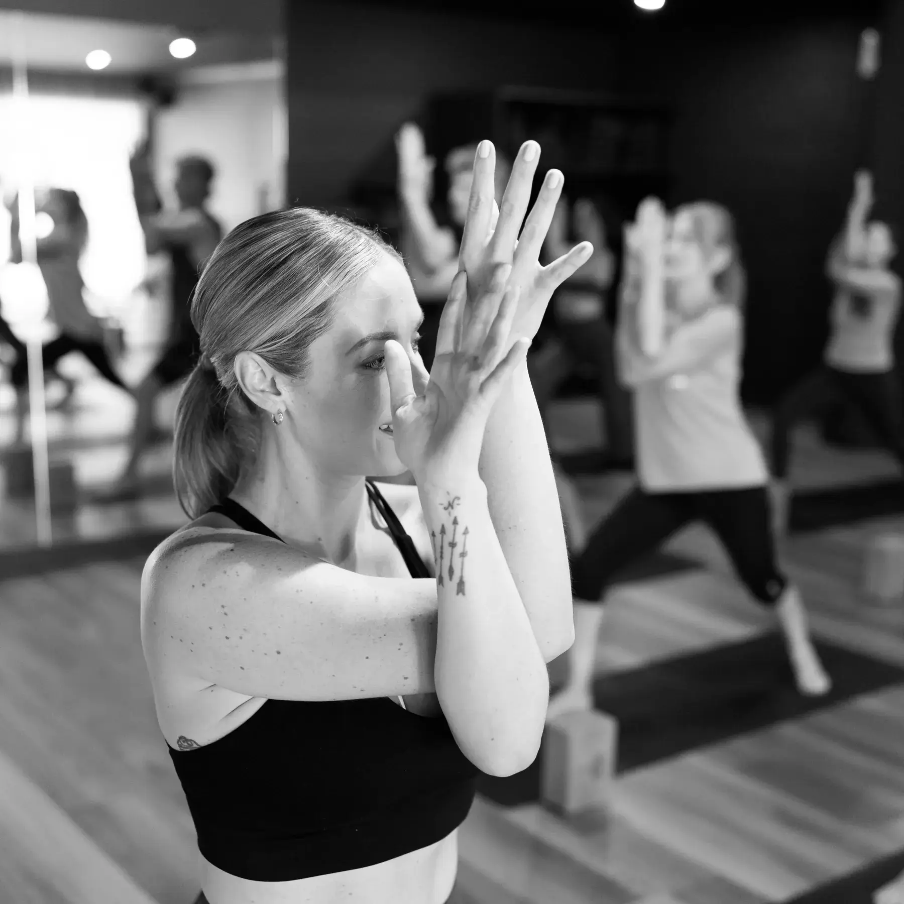 Black and white photo of a member in a deep twist with her hands pressed together in prayer as the class practises behind her