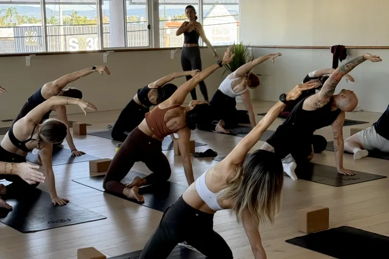 A class kneeling in a side stretch by the big windows of the Flolo Mermaid Beach studio as the teacher demonstrates